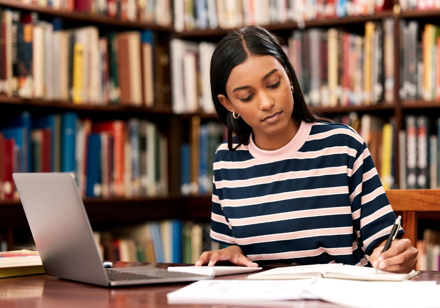 Jovem adulta estudando na biblioteca. Sobre uma mesa há notebook, cadernos e livros abertos. Ela está fazendo anotações em um caderno