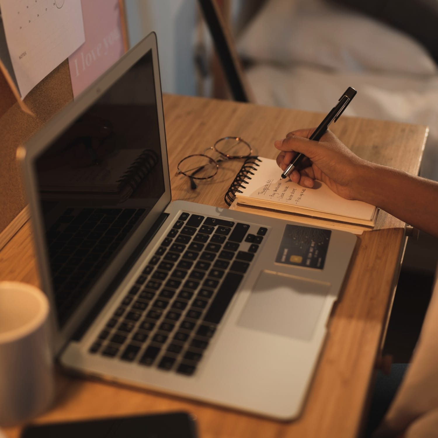 imagem de um notebook sobre uma mesa e uma pessoa escrevendo em um caderno ao lado
